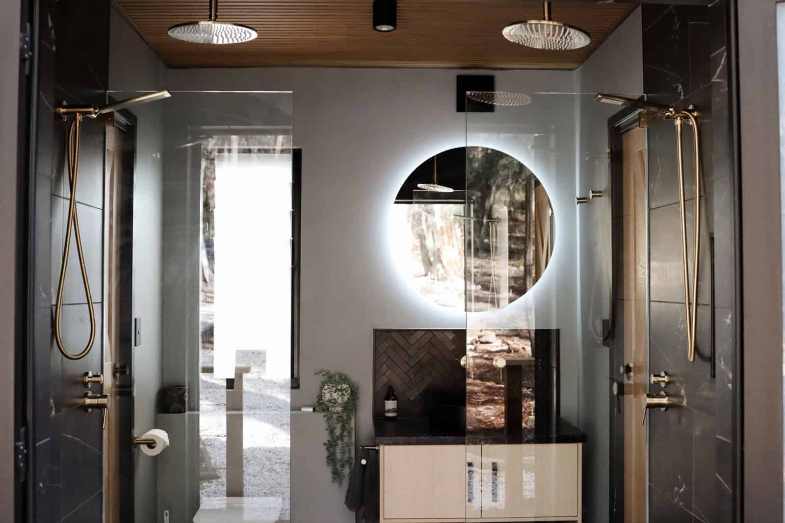 The SHAK's double shower with twin brass rainfall heads, dark marble tile, a timber ceiling, and a round backlit mirror above a timber vanity