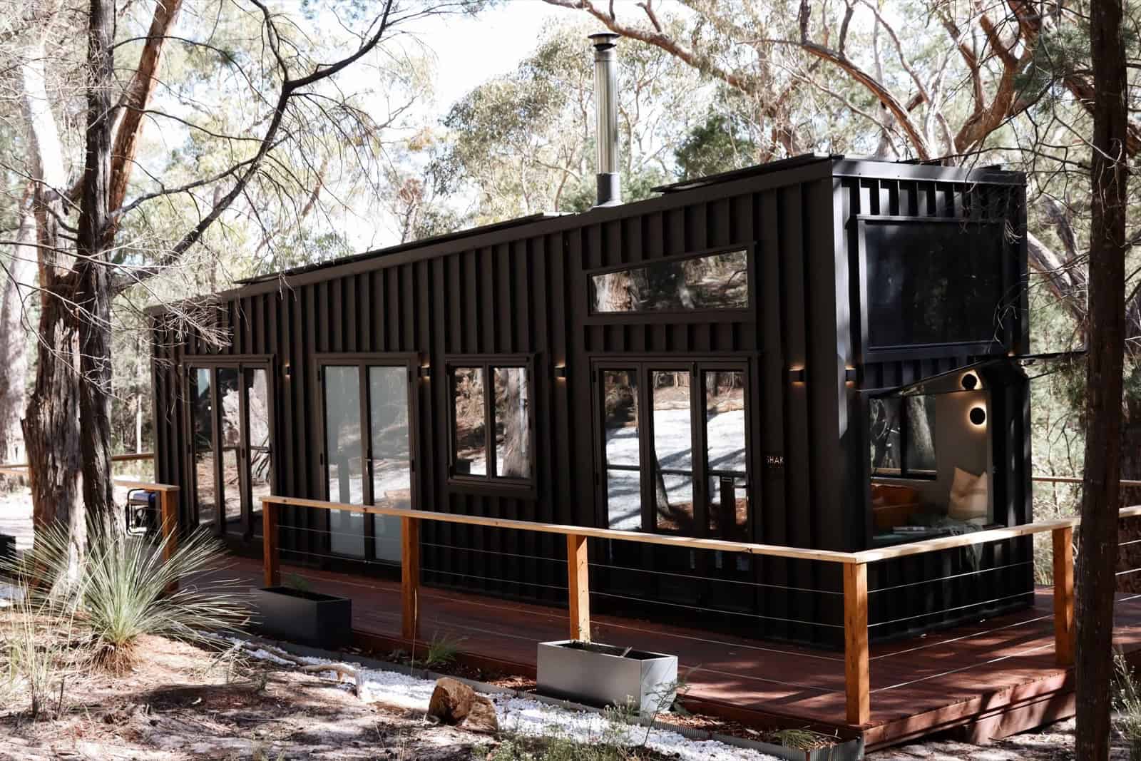 The SHAK by Removed Tiny Homes, a dark sculptural off-grid tiny house with an angular roofline and a wood-fire flue, set on a timber deck among the gum trees of the Grampians