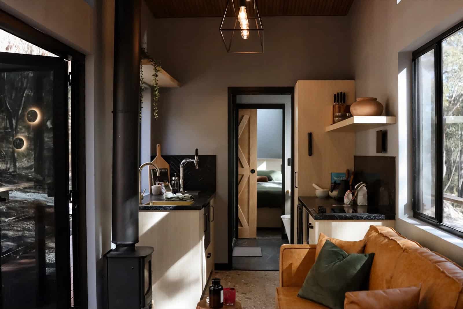 The SHAK kitchen with a dark stone benchtop, timber cabinetry, brass tapware and wine on tap, a herringbone-tile splashback, and a black wood-burning fireplace beside a leather sofa