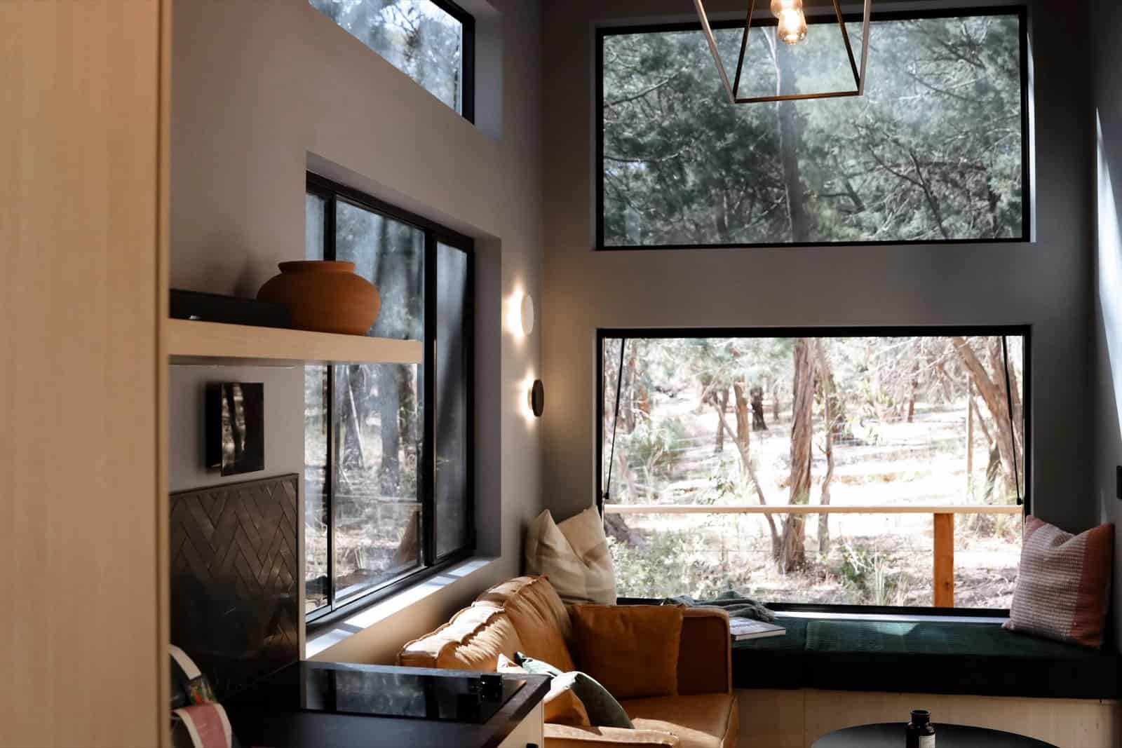 The SHAK living area with a tan leather sofa and a green velvet window seat beneath stacked windows framing the bush, with a brass pendant and timber shelving
