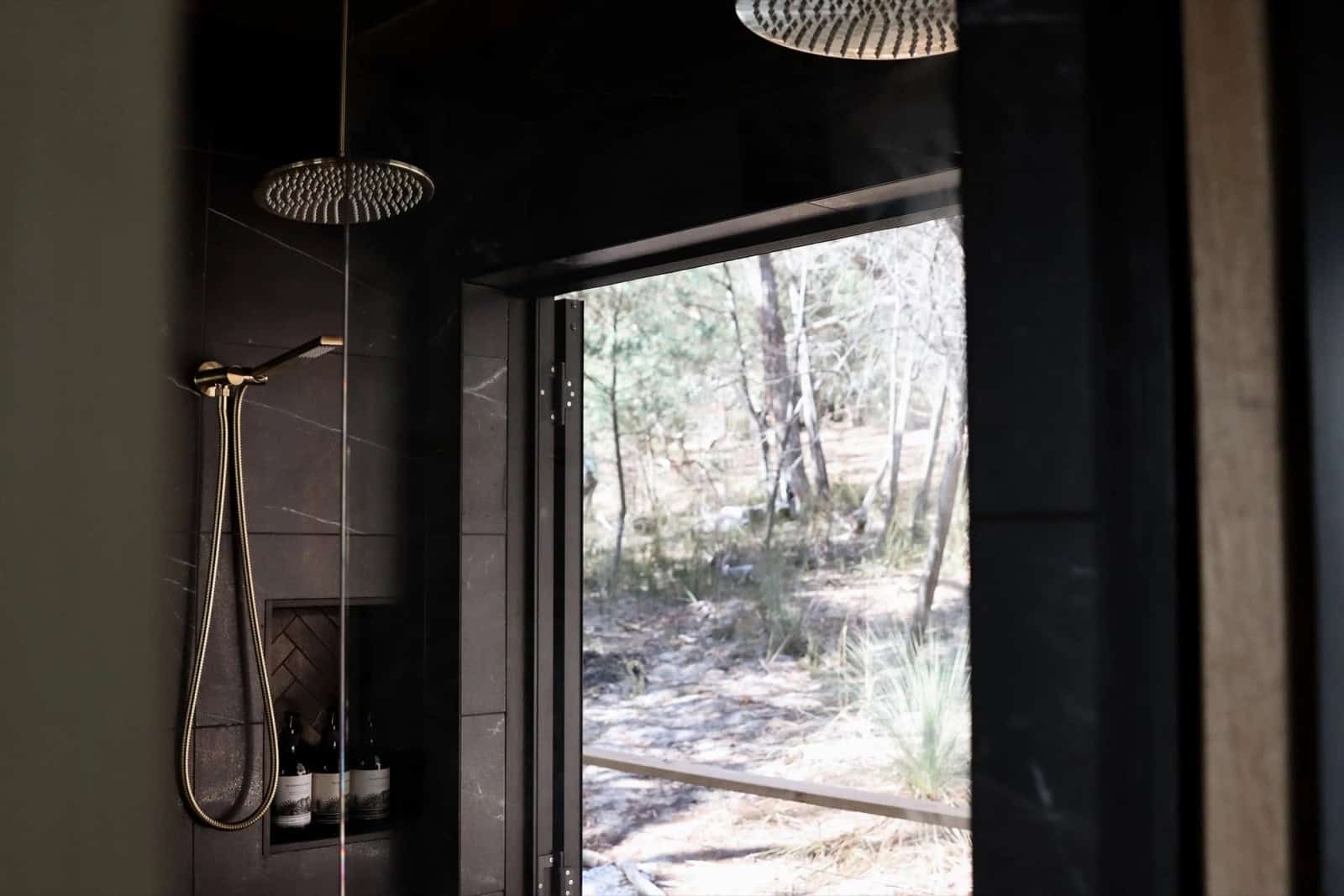 A brass rainfall shower in the SHAK with a full-height glass door open directly onto the surrounding bushland, creating an open-air shower