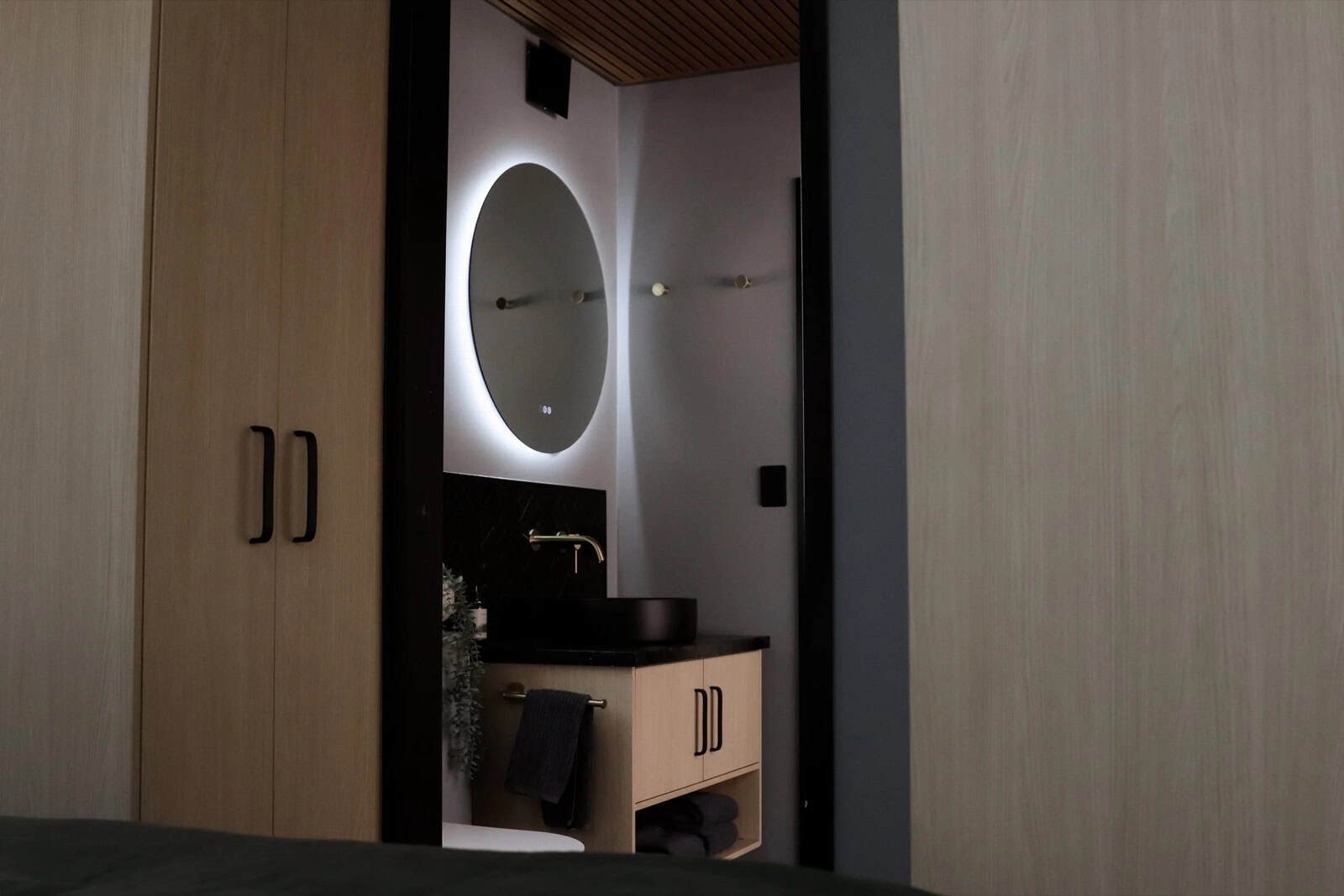 The SHAK bathroom vanity with a black stone top, black vessel basin, brass wall-mounted tap, and a round backlit mirror, framed by timber wardrobe joinery