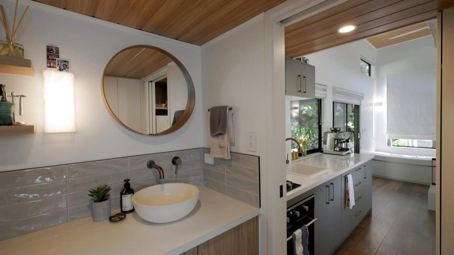 The Springbrook bathroom with a timber vanity, a stone vessel basin, a round timber-framed mirror, brushed-brass tap, and glossy grey tile, opening back toward the kitchen