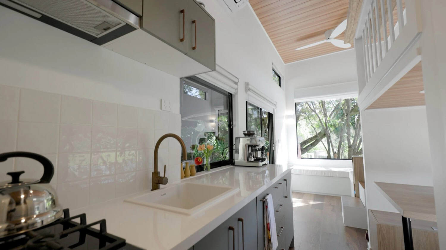 The interior of the Springbrook 7.2 by Removed Tiny Homes, with a sage-green galley kitchen, a window-seat nook, a timber-lined ceiling, and a corner stairway up to the loft
