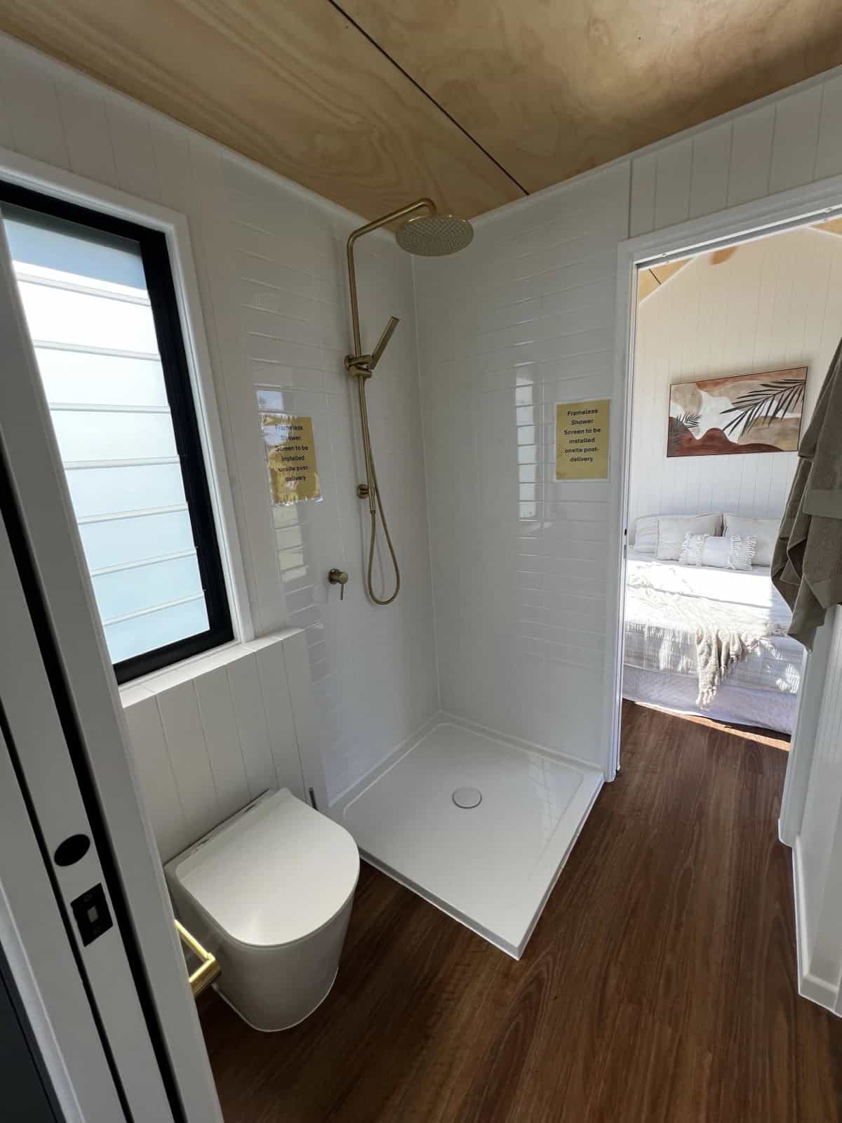 The Tallebudgera ensuite with a white-tiled shower, brushed-brass rainfall head, a modern toilet, and a louvre window, opening through to the bedroom