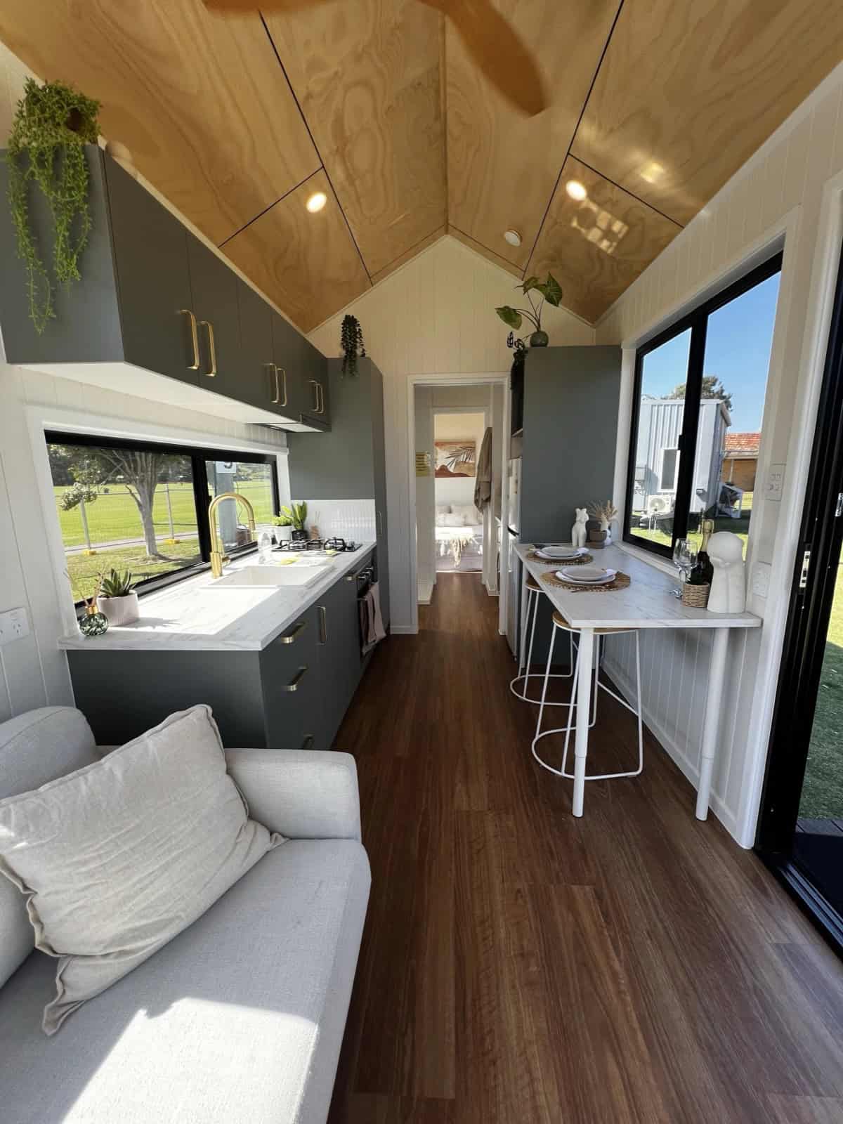 Interior of the Tallebudgera looking down its length, with a charcoal galley kitchen on the left, a breakfast bar on the right, a vaulted timber ceiling, and the bedroom doorway at the far end