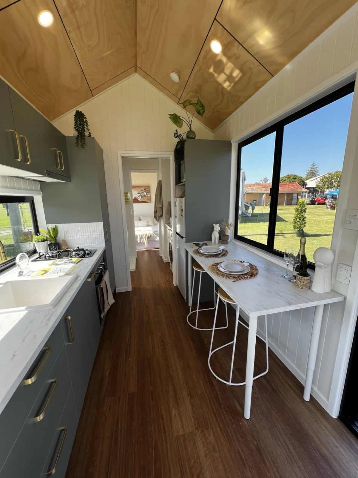 The Tallebudgera kitchen with charcoal cabinetry, brass handles and tap, a white benchtop and splashback, a cooktop and oven, and a marble-look breakfast bar beneath a large picture window