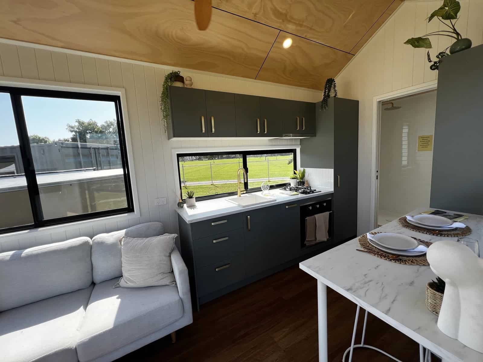 The open-plan living and kitchen of the Tallebudgera with a grey sofa, charcoal cabinetry, a white benchtop, brass tapware, and a marble-look dining table