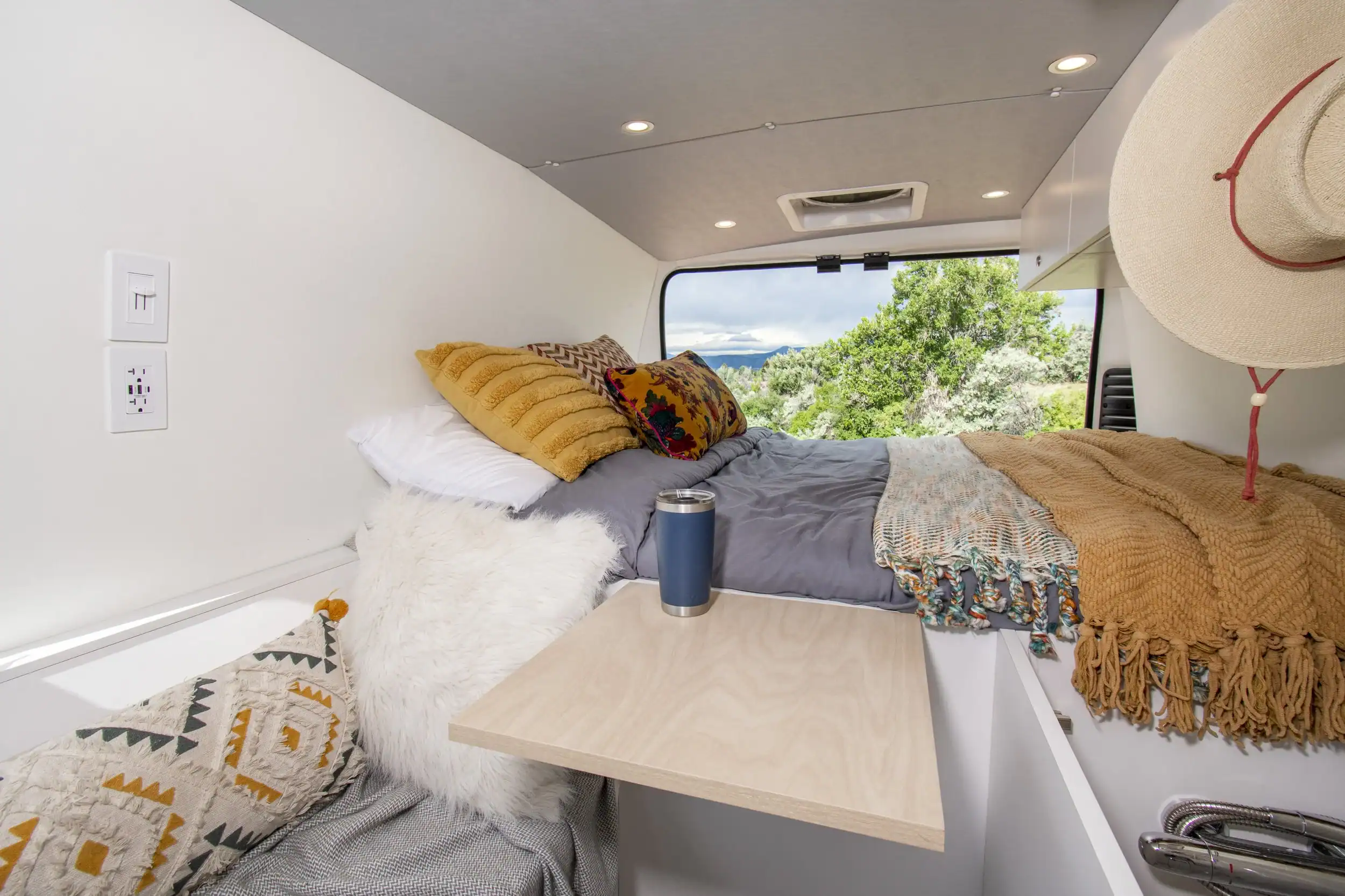 Interior of a tiny house bedroom featuring a bed with colorful pillows and a view of green trees out.