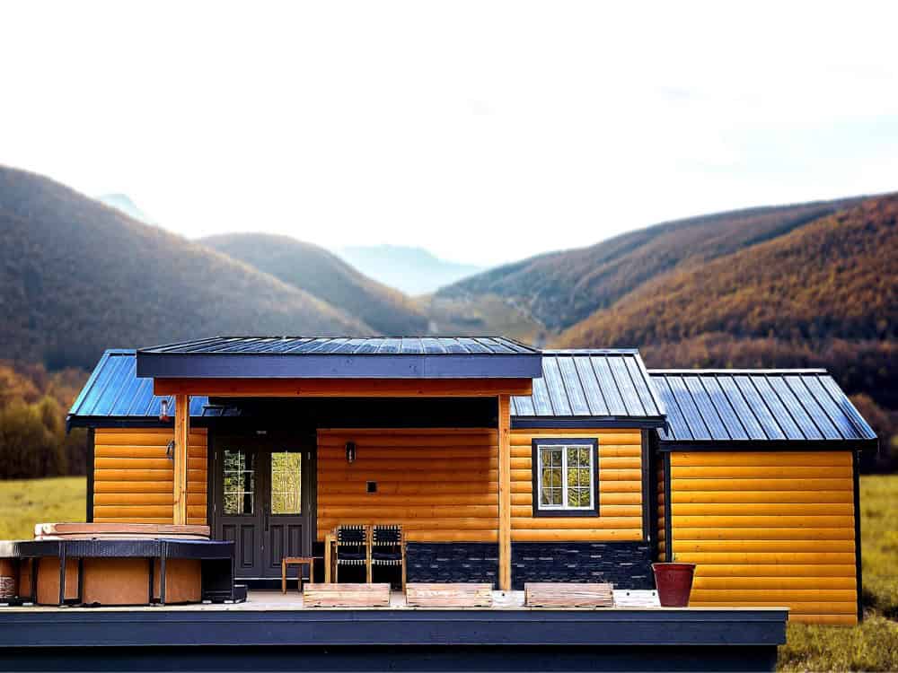 Rolling Bear Black Bear community cottage with log siding, stone skirting, dark metal roofs, a covered porch, and a hot tub, set against autumn mountains