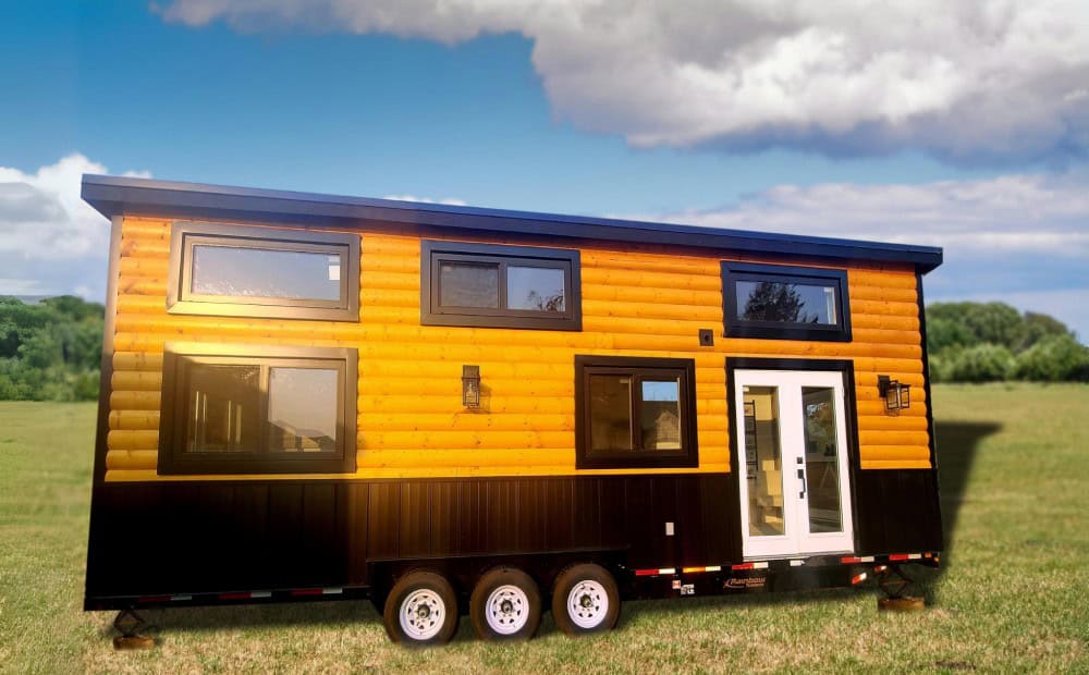 Rolling Bear Cinnamon Bear family lodge in a meadow, with log siding, a black base, dormer windows, French doors, and a tri-axle trailer