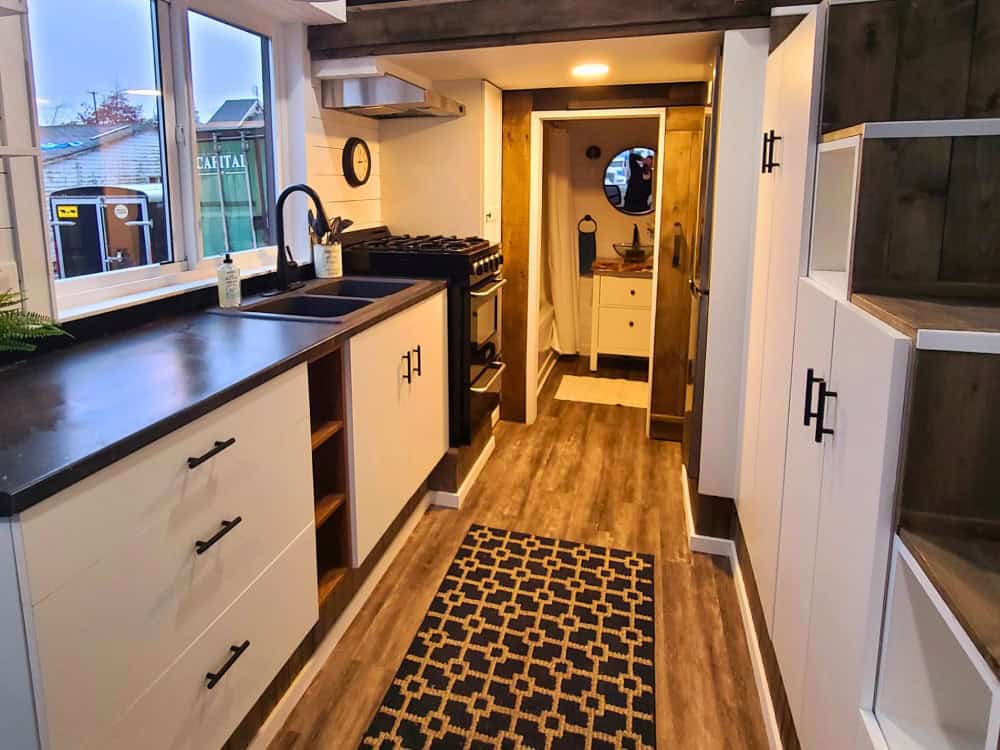 Koala Bear galley kitchen with a gas range, double-basin sink, white shaker cabinets, live-edge shelving, and a hallway to the bathroom