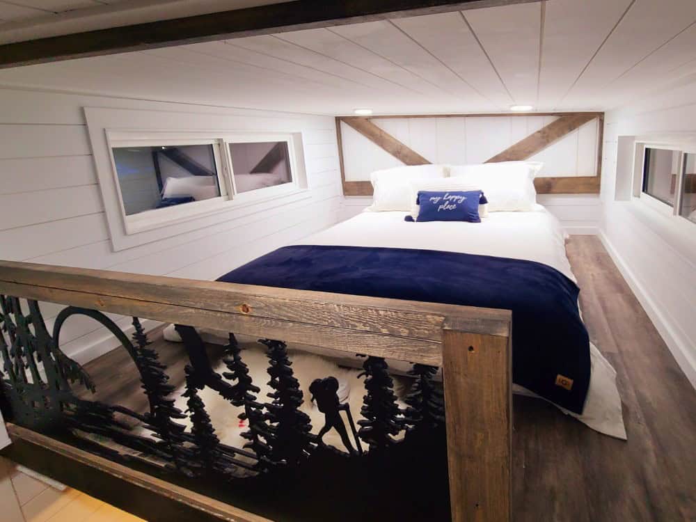 Queen lofted bedroom in the Koala Bear with white shiplap, a reclaimed-wood headboard, dormer windows, and a custom metal railing of a hiker in a pine forest