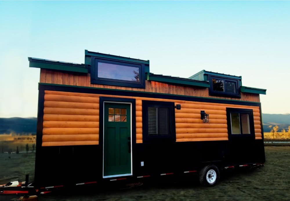 Rolling Bear Salmon Bear tiny house on wheels at dusk, with log siding, a green door, two dormers, and mountains behind