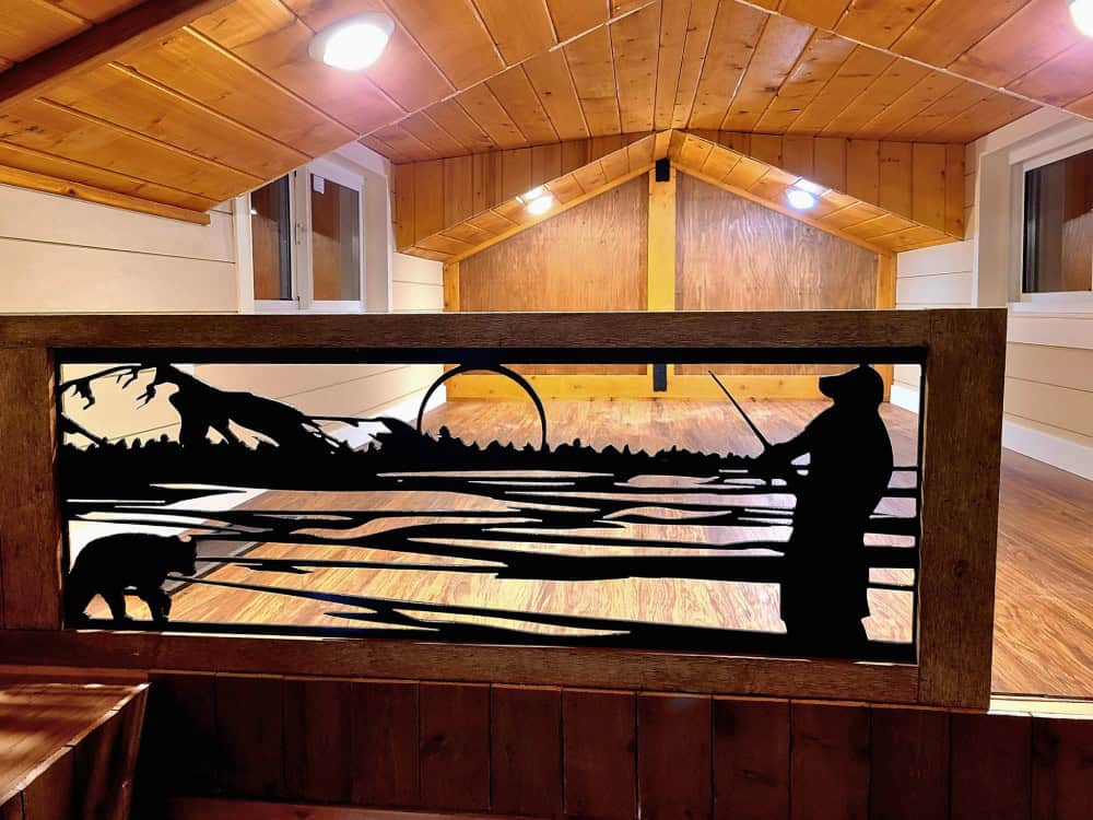 Custom laser-cut metal loft railing in the Salmon Bear depicting a fly fisher and a bear catching salmon, under a vaulted cedar ceiling