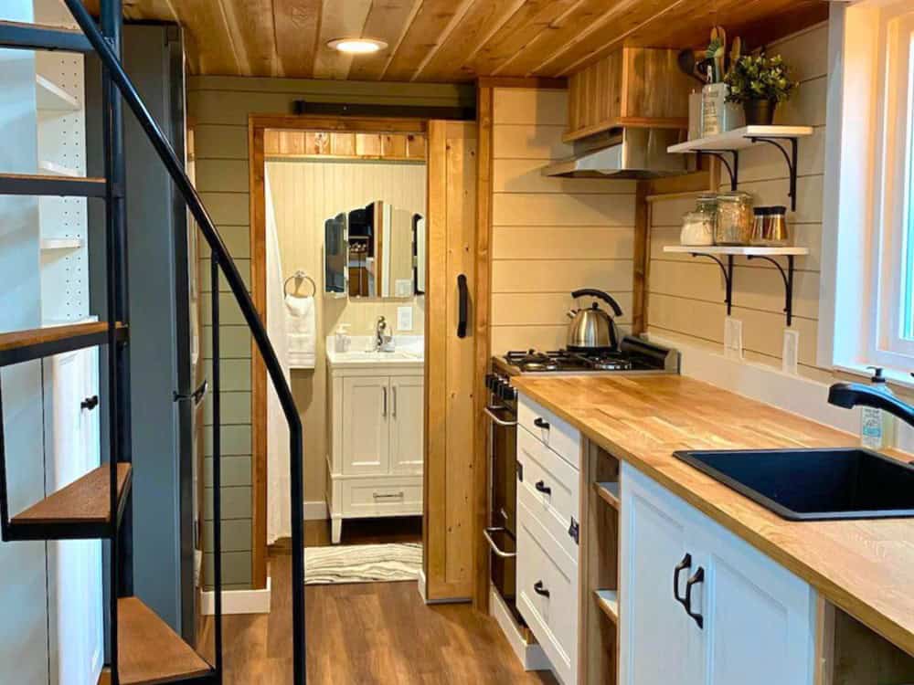 Elevated living room in the Skylar Bear with a black sofa, wood stove, reclaimed wood-tile accent wall, and vaulted cedar ceiling above the kitchen