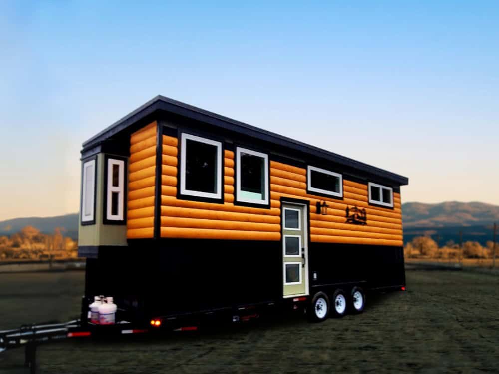 Rolling Bear Skylar Bear tiny house on wheels at dusk, with log siding, a black base, a cream accent end wall, and a tri-axle trailer