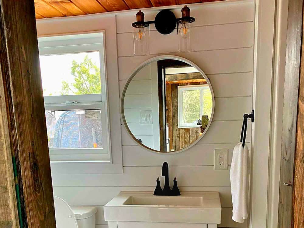 Deluxe bathroom in the Spirit Bear with white shiplap walls, a vanity, a round mirror, Edison-style sconces, and a window