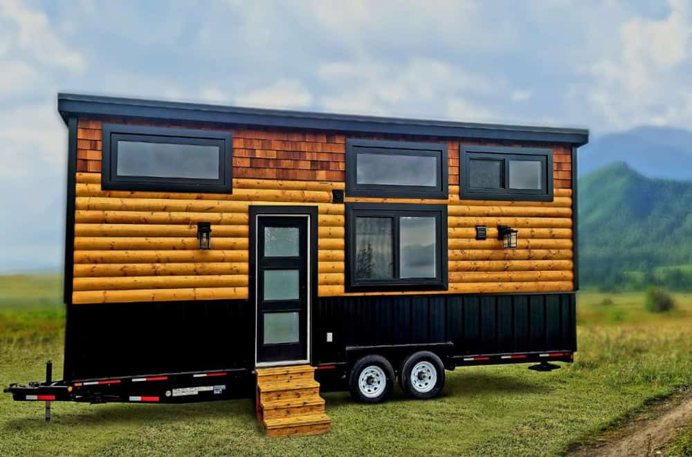 Rolling Bear Spirit Bear tiny house on wheels in a mountain meadow, with log siding, cedar-shake upper, a black base, a tall glass door, and dormer windows