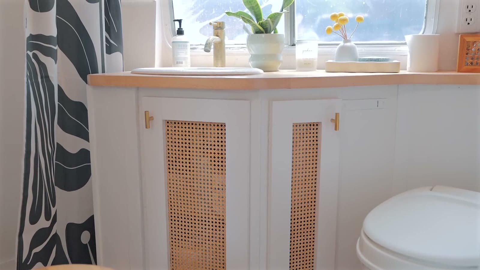 Bathroom in a renovated Avion trailer with an oval sink set in a wood counter, cane-front cabinets with brass pulls, a black-and-white floral shower curtain, plants on the windowsill and a white toilet