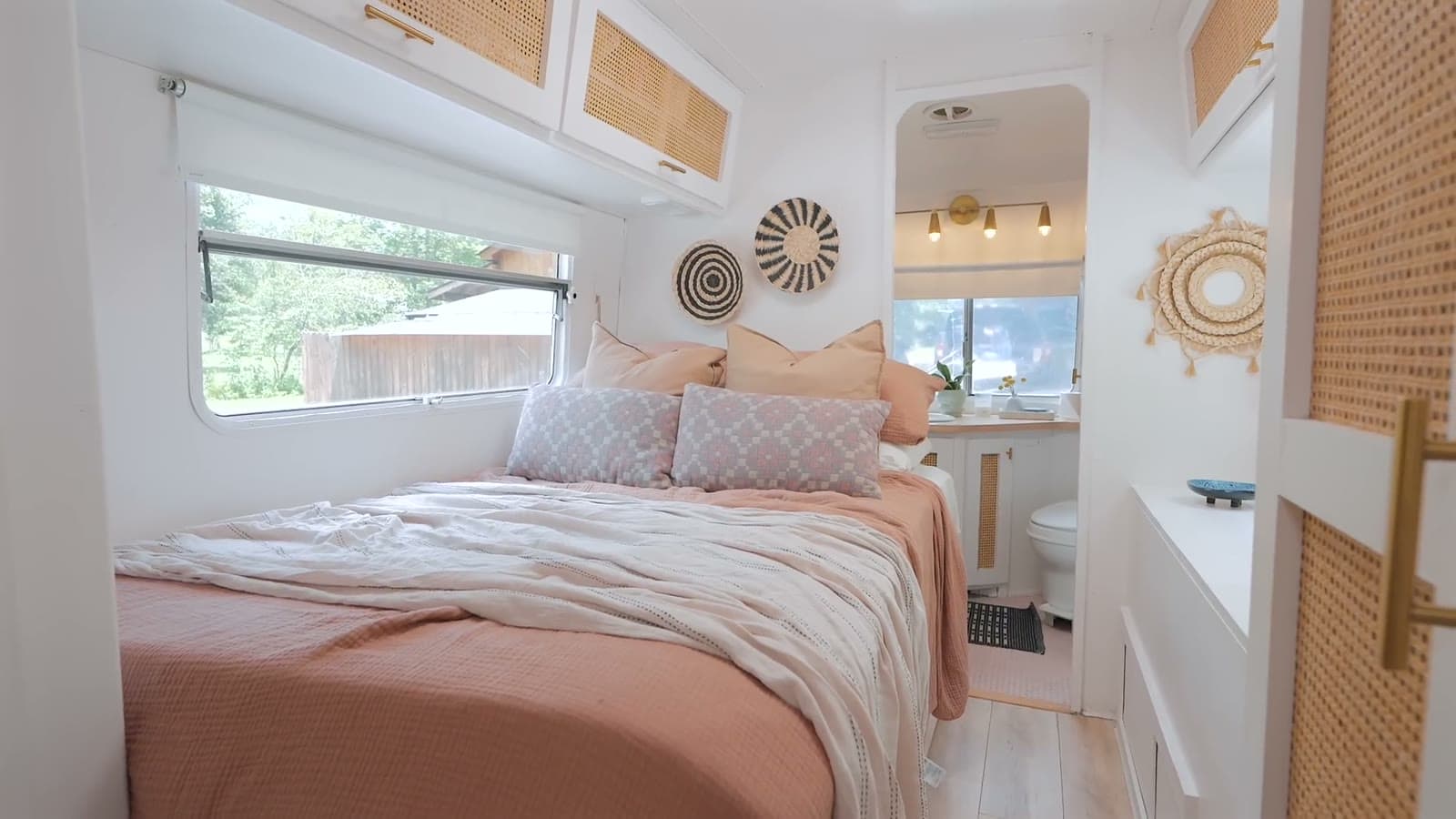 Bedroom in a renovated Avion trailer with a queen bed in terracotta and cream bedding, woven wall art, cane cabinet fronts, windows on both sides and the bathroom door beyond