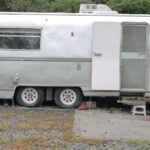 Silver 1973 Avion La Grande travel trailer parked on gravel among trees, rounded aluminum body with awning windows, entry door open, roof air conditioner and a wooden step chest at the door
