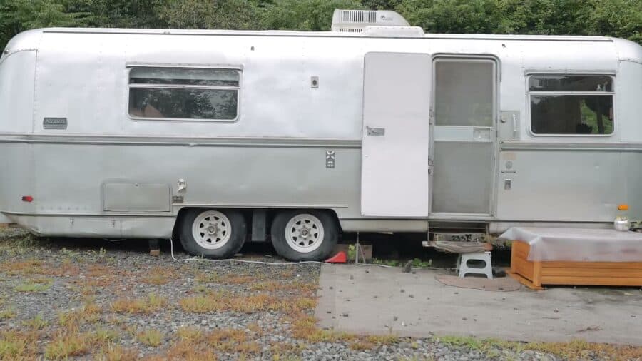 Silver 1973 Avion La Grande travel trailer parked on gravel among trees, rounded aluminum body with awning windows, entry door open, roof air conditioner and a wooden step chest at the door