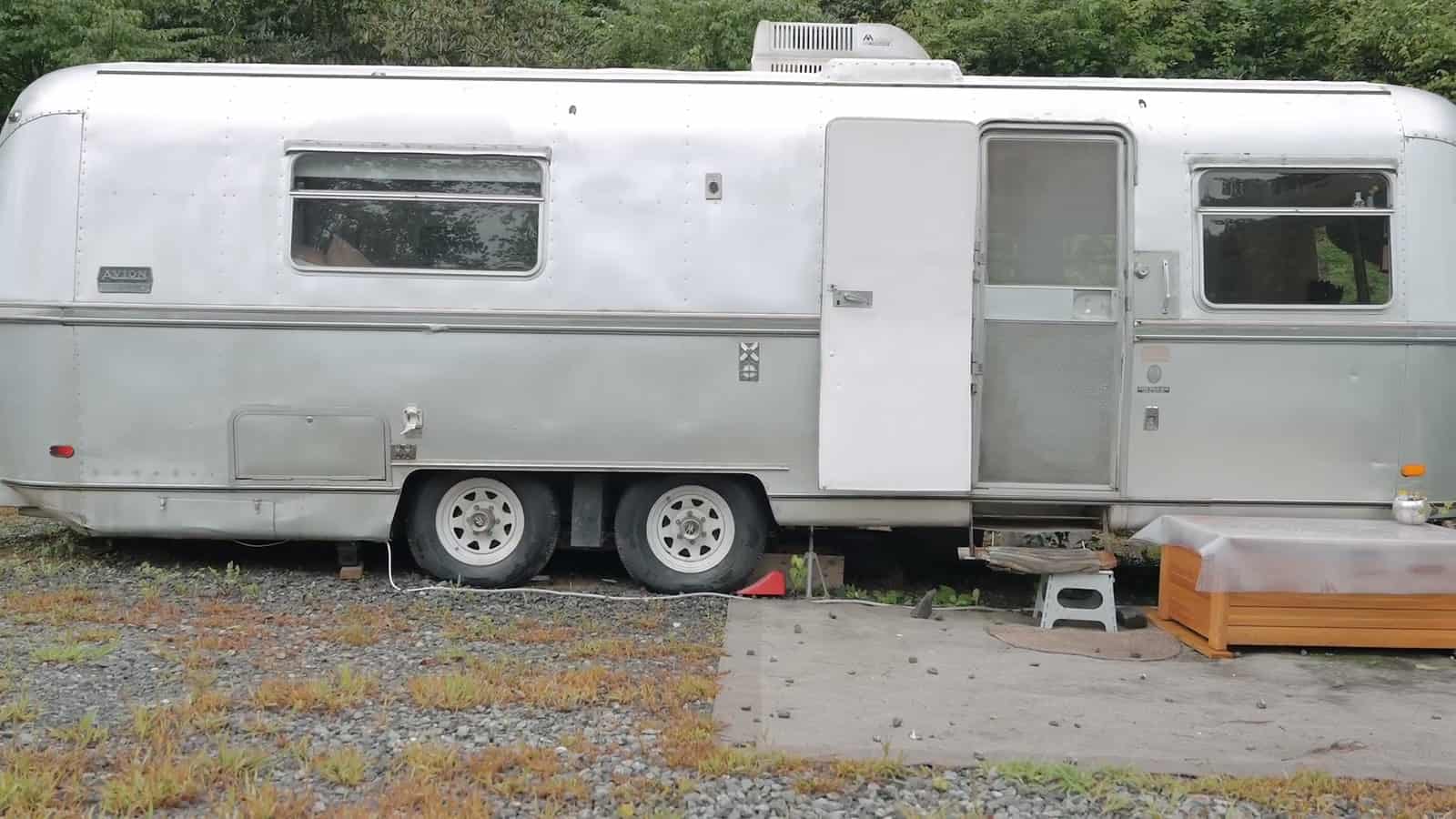Silver 1973 Avion La Grande travel trailer parked on gravel among trees, rounded aluminum body with awning windows, entry door open, roof air conditioner and a wooden step chest at the door