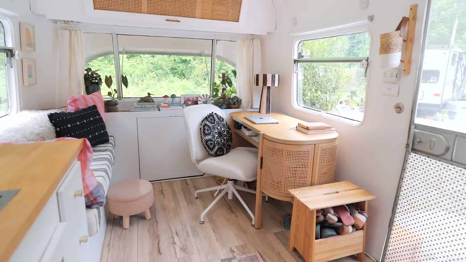 Living lounge inside a renovated Avion trailer with wraparound windows, a striped bench with pillows, pale pink ottoman, cane-front wood desk with a white chair, and a slatted wood lamp