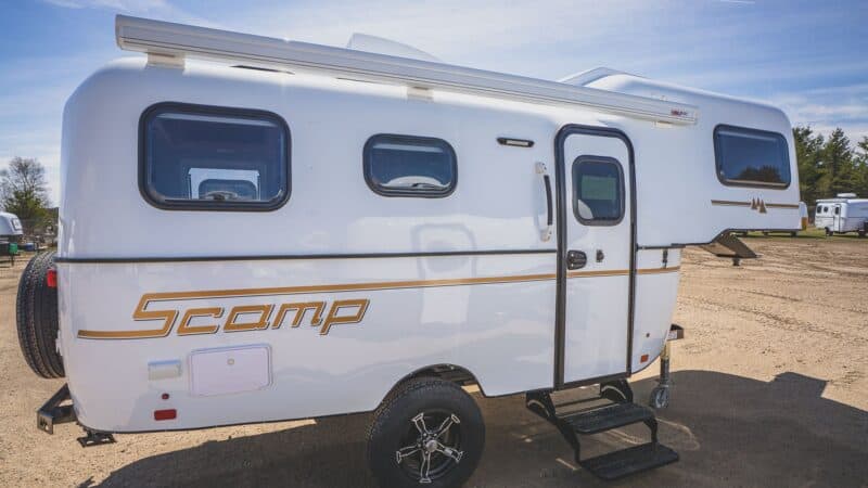 White Scamp 19 fifth wheel with gold stripes parked on a gravel lot