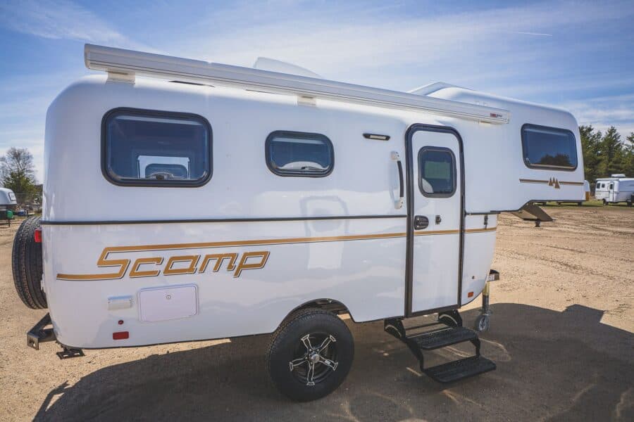 White Scamp 19 fifth wheel with gold stripes parked on a gravel lot