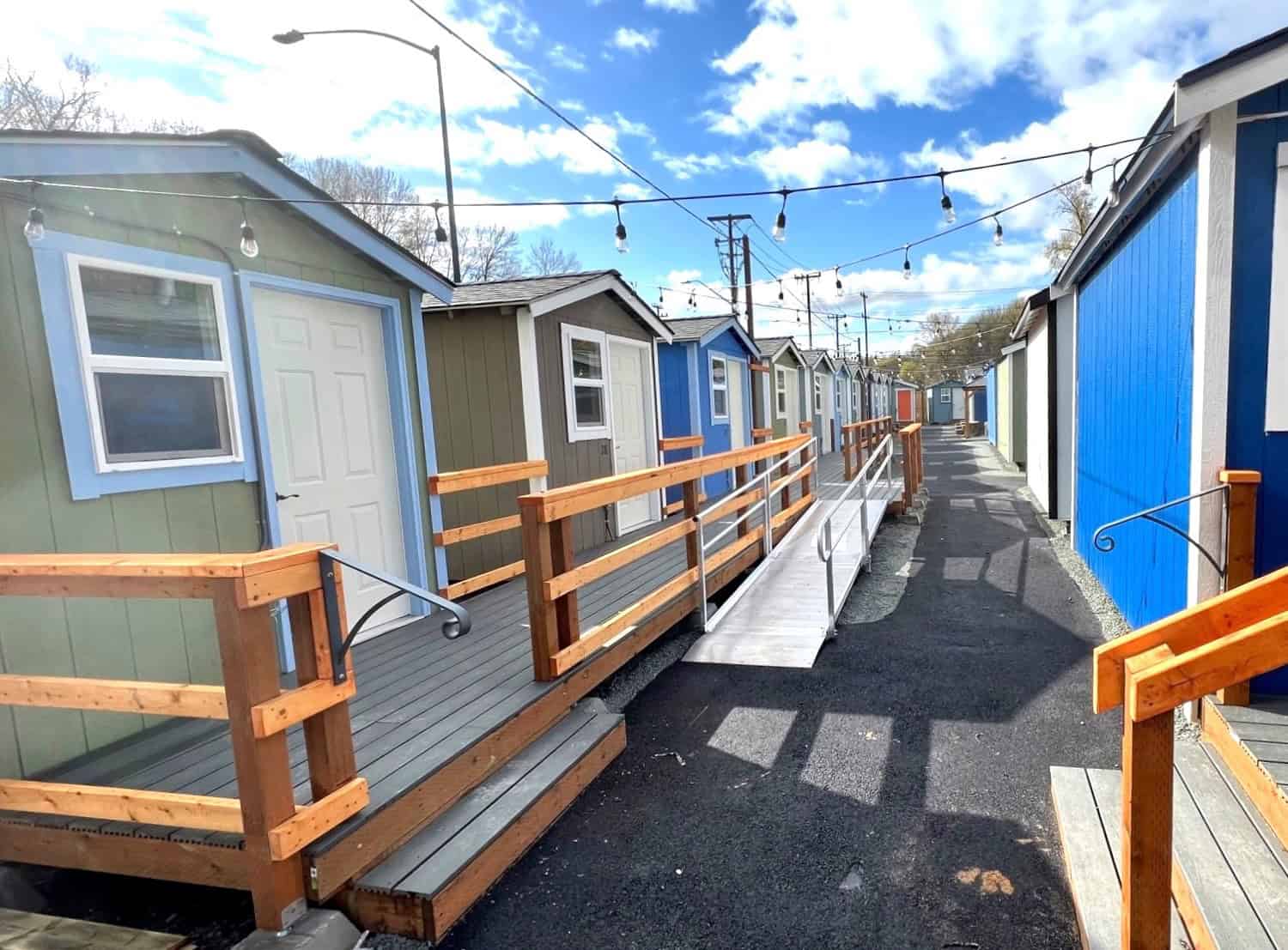 Cedar boardwalk and accessible ramp running past a row of green and blue tiny houses under string lights at a Seattle village