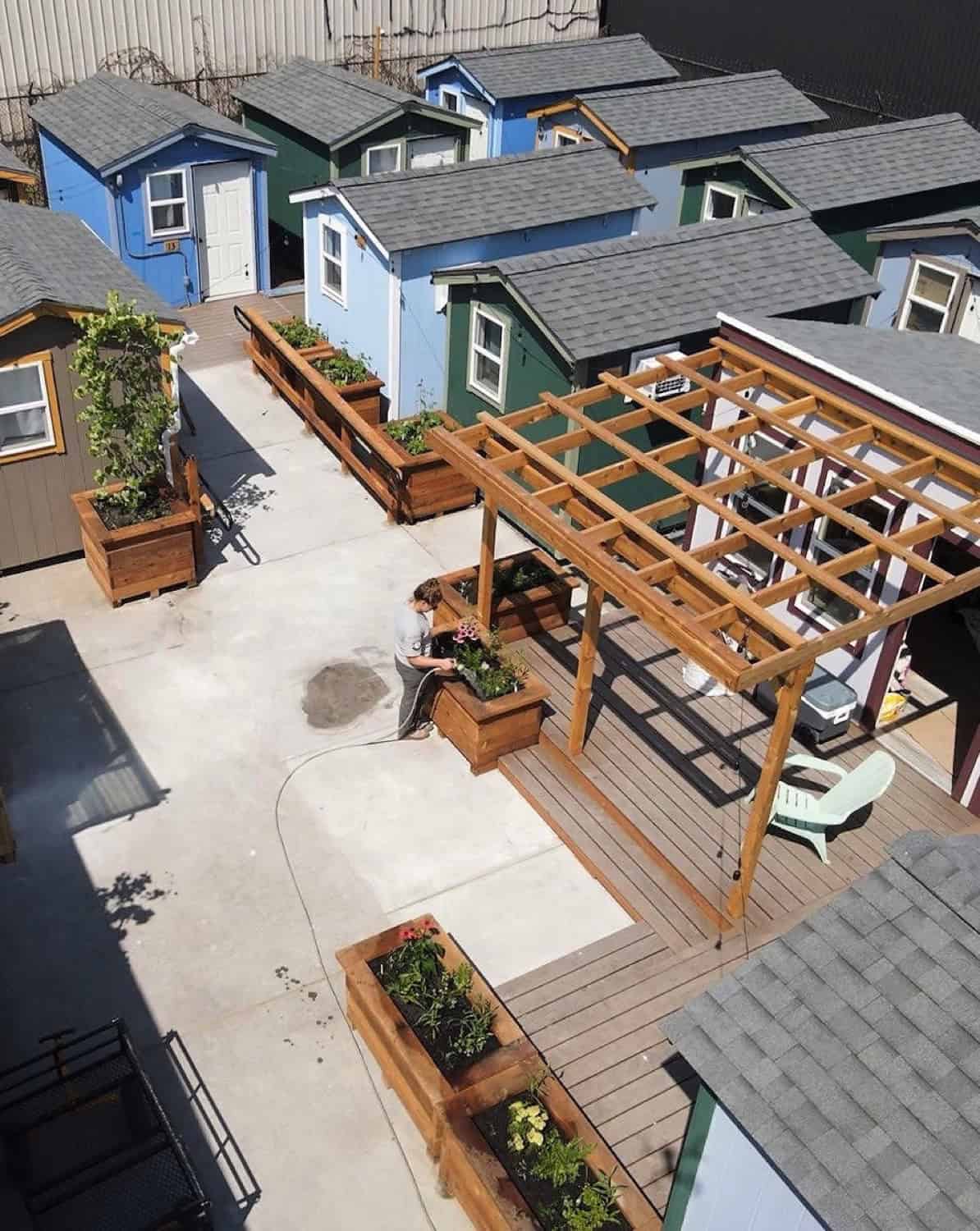 Aerial view of a Seattle tiny house village showing shingled gable roofs, a cedar pergola over a deck and raised planter beds full of flowers