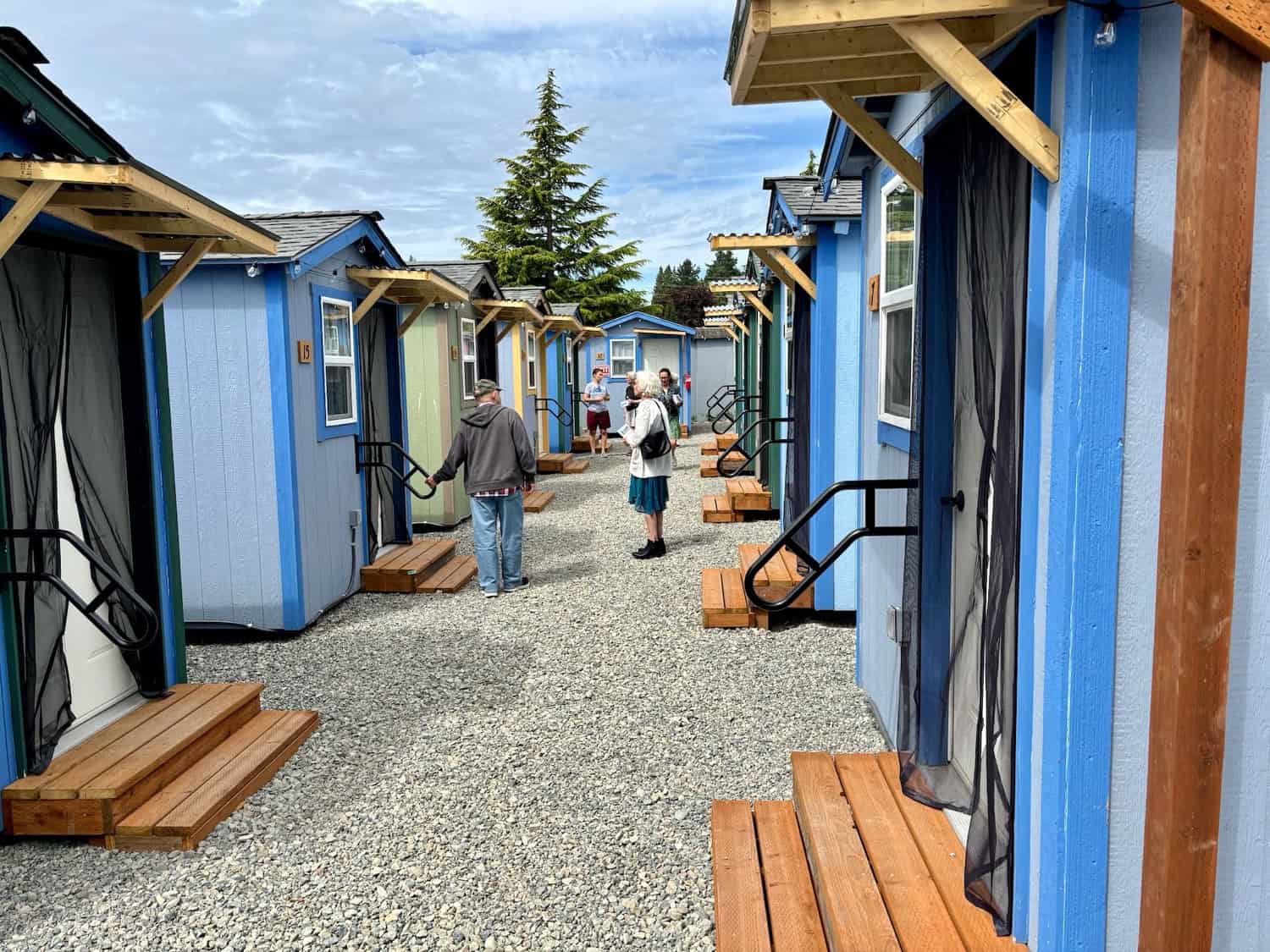 Residents walking a gravel lane between blue and green tiny houses with cedar porch awnings, handrails and numbered doors at a Seattle village