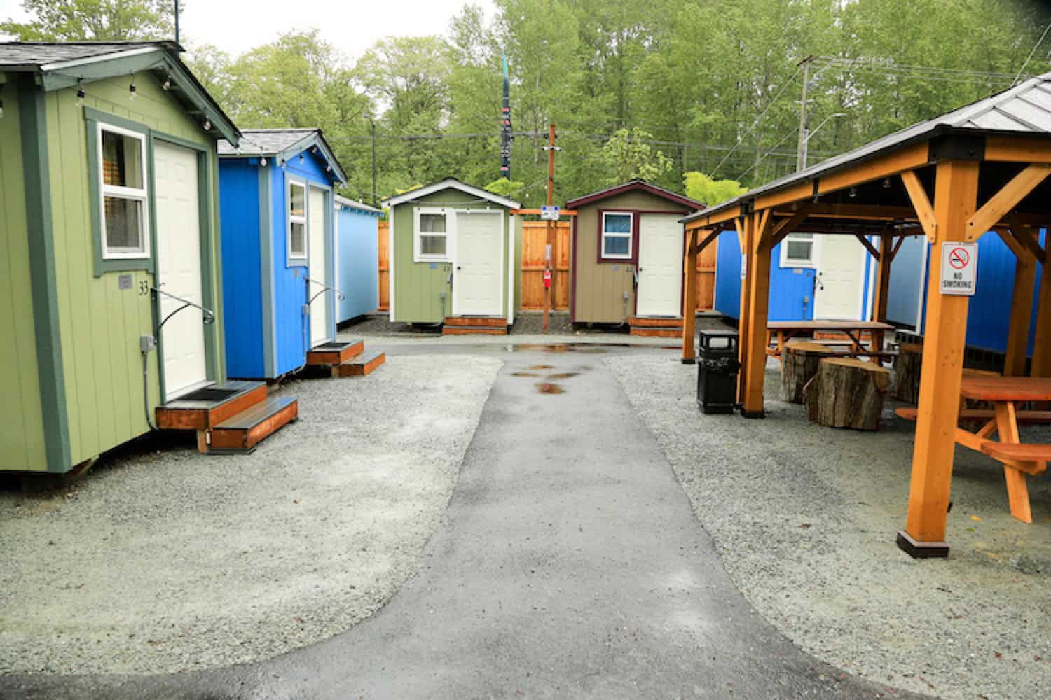 Green, blue and tan tiny houses around a gravel courtyard with a timber gazebo, picnic tables and log stools at a Seattle village