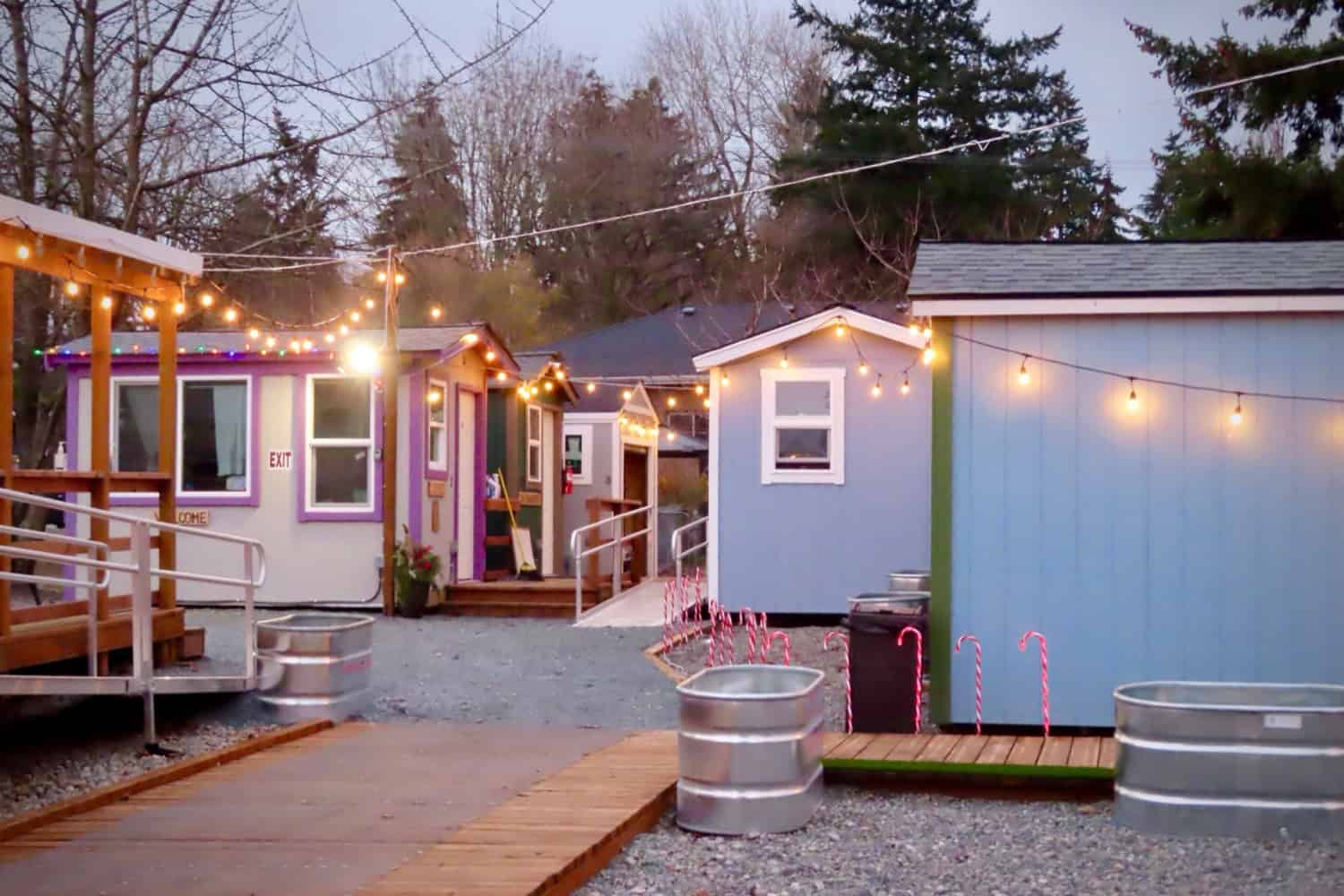 Tiny houses in purple, blue and tan glowing under string lights at dusk with holiday decorations at a Seattle village