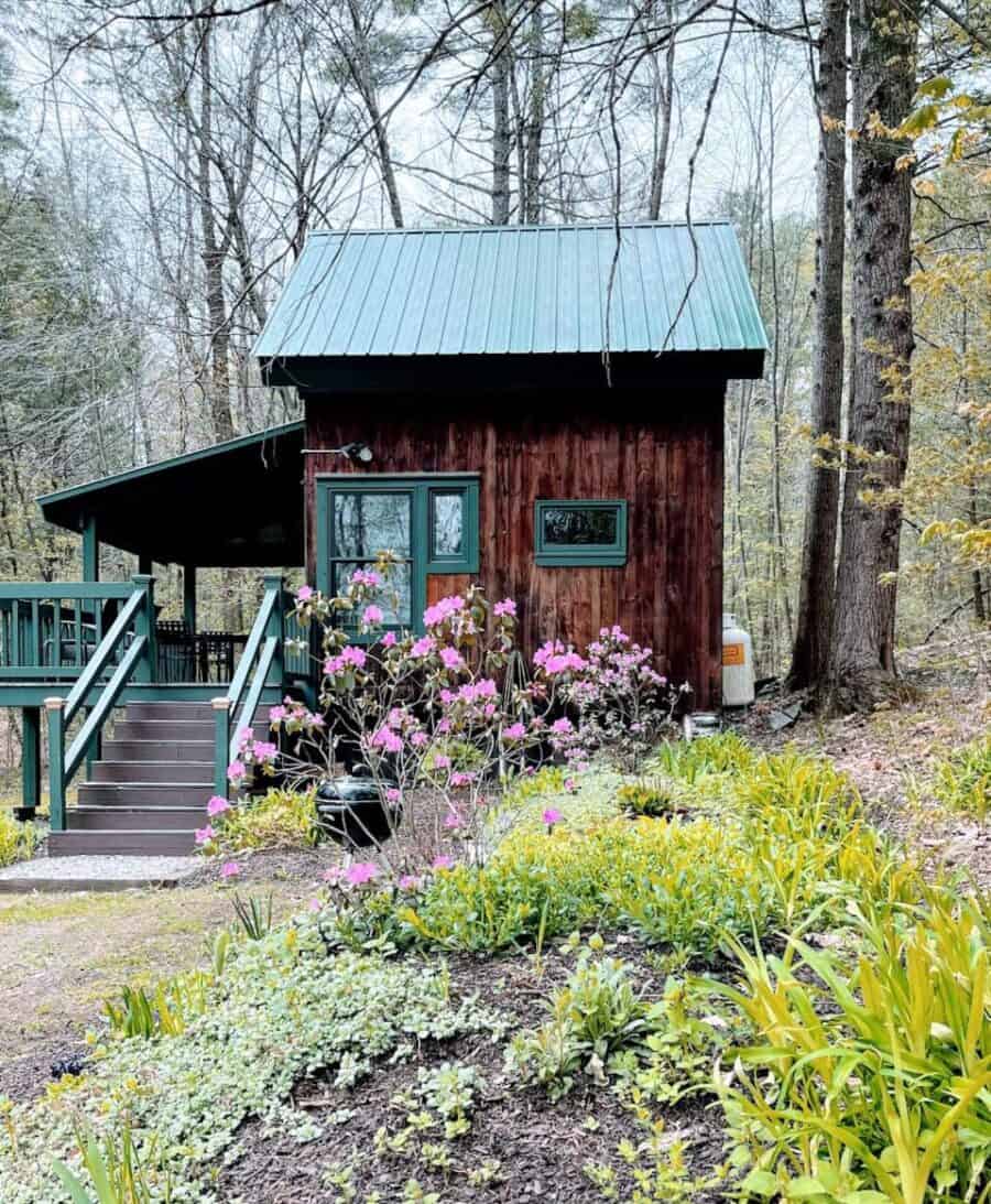 Rustic tiny house with a green metal roof surrounded by blooming flowers and trees in Vermont.
