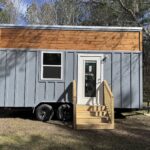 Gray board-and-batten Shanti tiny house on a dual-axle trailer with a cedar accent band, shed roof, glass door, and wood steps in a wooded yard