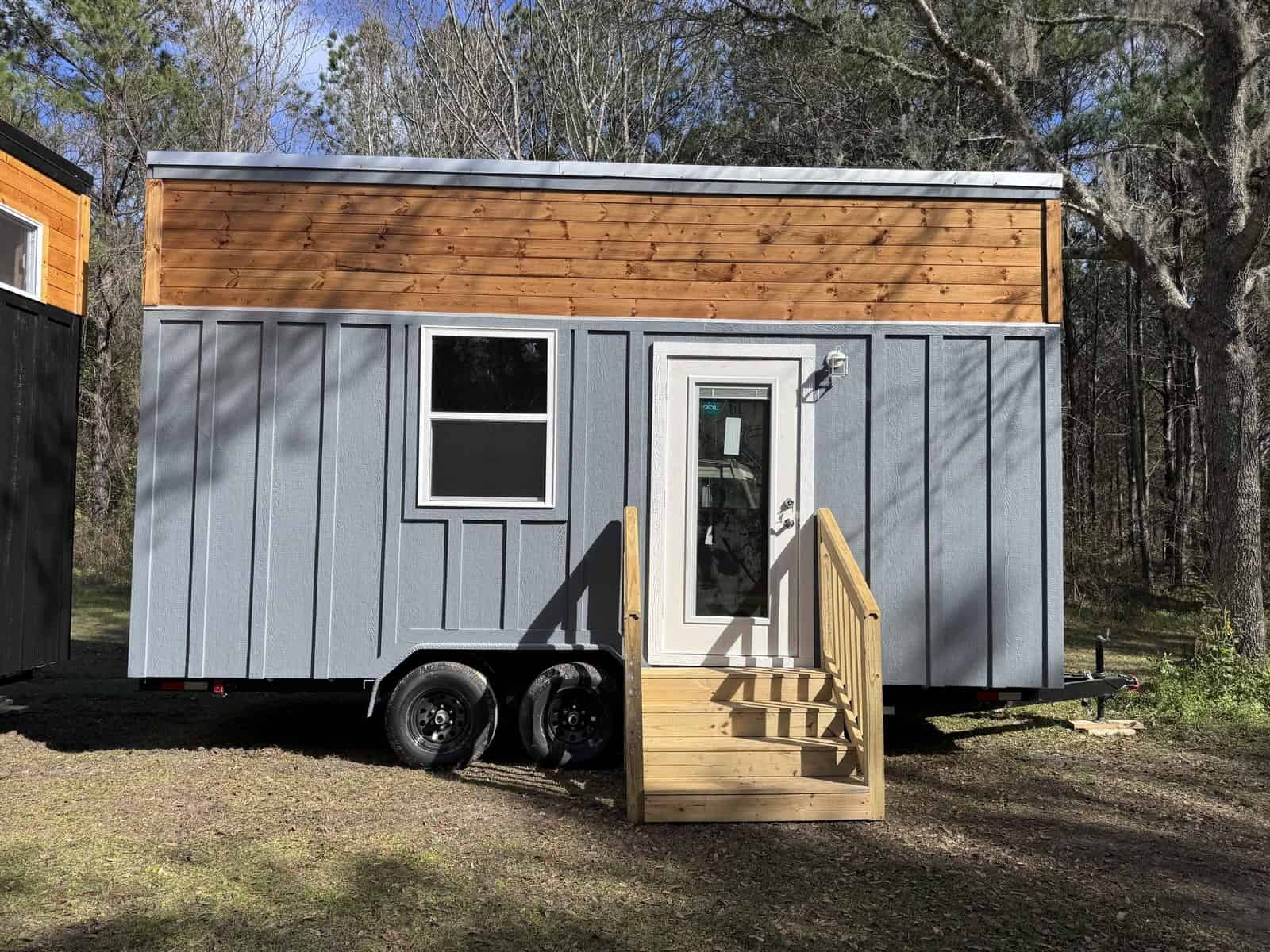 Gray board-and-batten Shanti tiny house on a dual-axle trailer with a cedar accent band, shed roof, glass door, and wood steps in a wooded yard