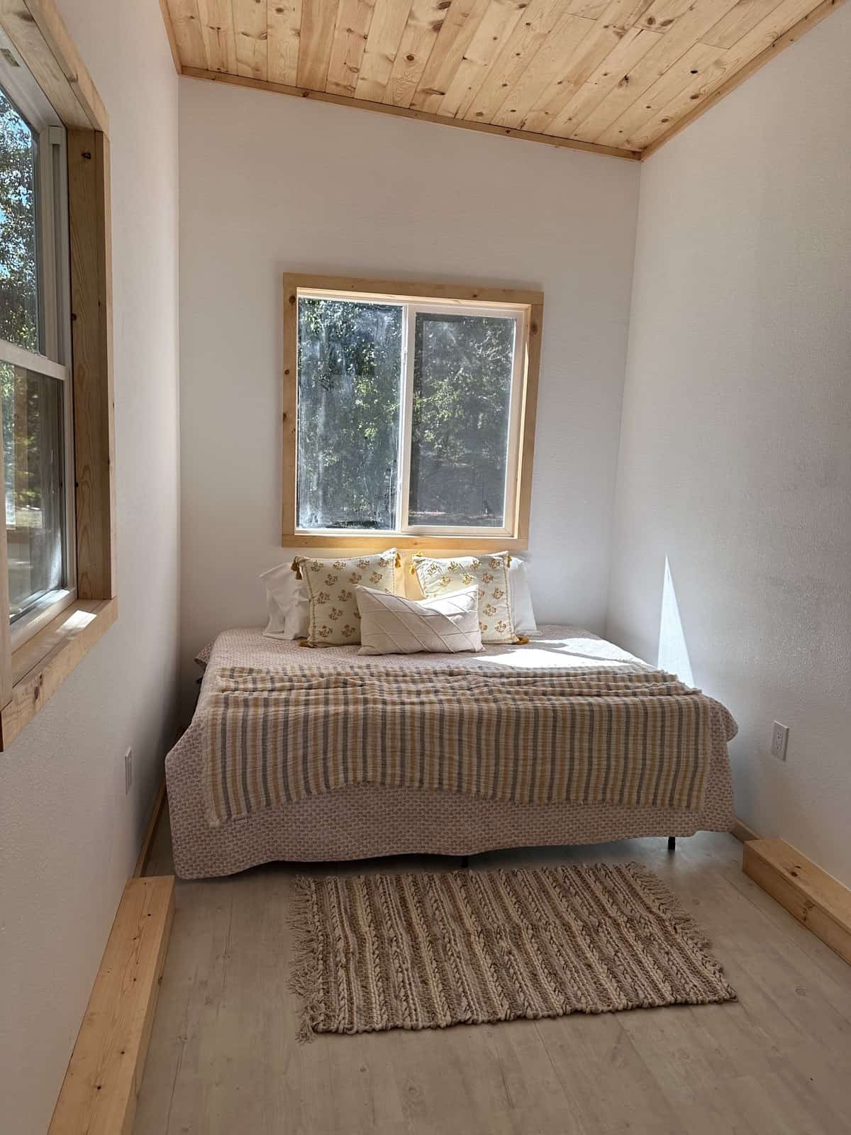 Ground-level queen bedroom in the Shanti tiny house with windows on two walls, a pine ceiling, and a woven rug