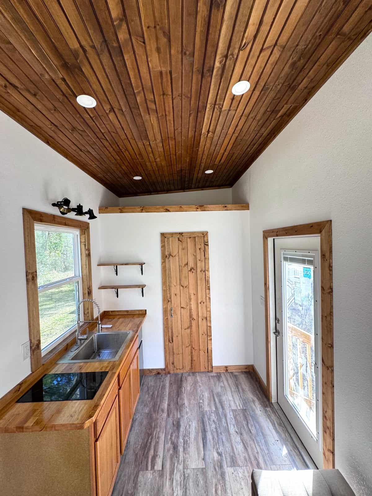 Interior of the Shanti tiny house with a stained pine ceiling, butcher-block kitchen, open shelving, a pine storage closet, and a glass entry door