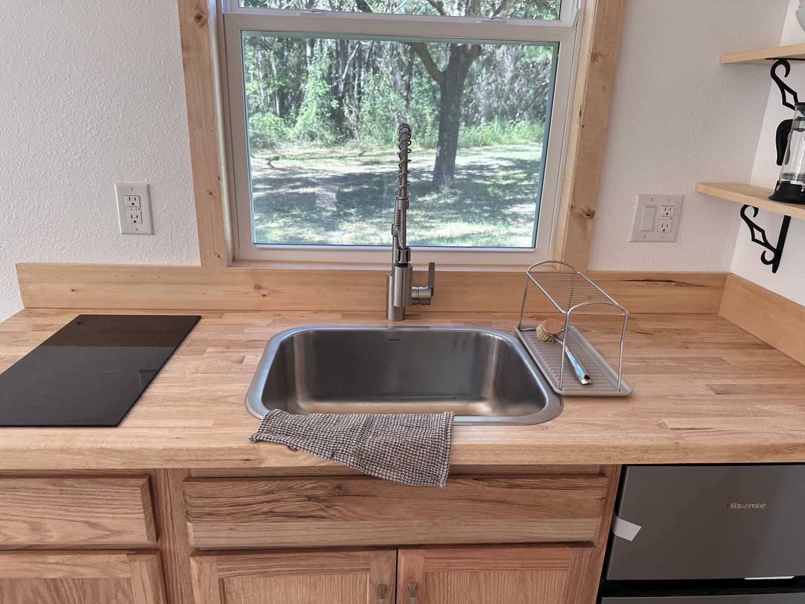Shanti tiny house kitchen detail with a butcher-block counter, stainless drop-in sink, gooseneck faucet, flush electric cooktop, and a window over the sink