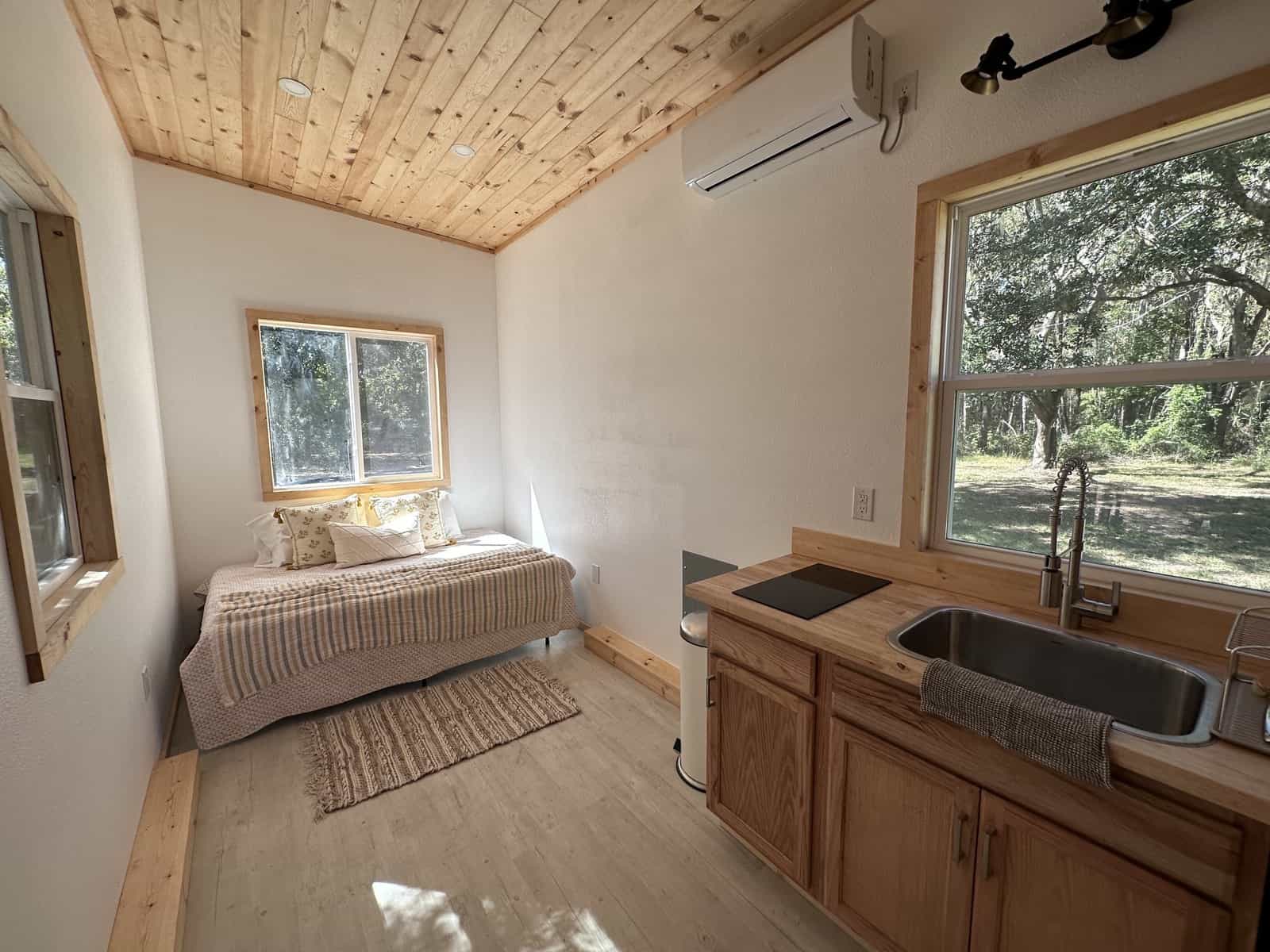Open floor plan of the Shanti tiny house with a ground-level queen bed at one end and a butcher-block kitchen with a mini-split and large windows at the other