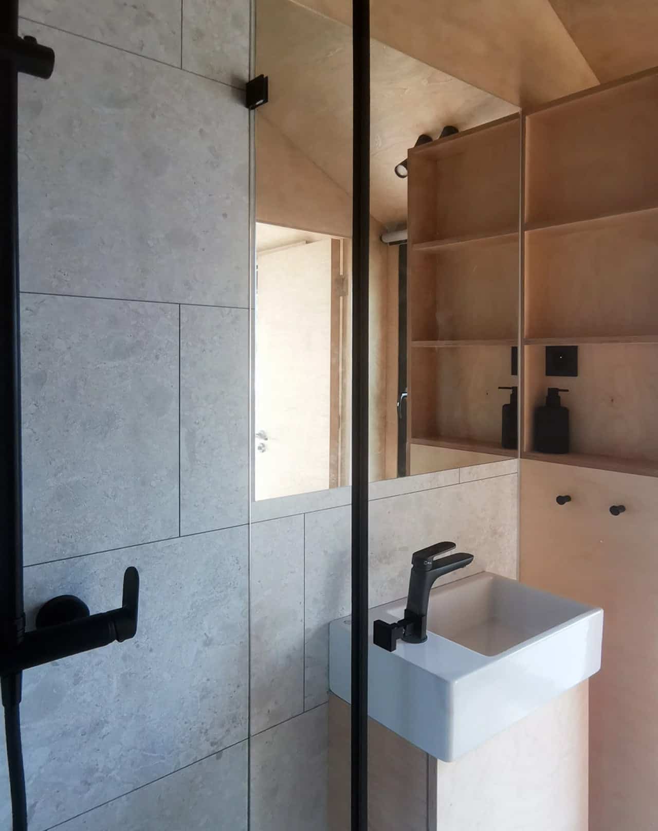 Bathroom in the Shoji tiny house with a large-format tiled walk-in shower behind a frameless glass partition, white wall-hung basin, black fixtures, and plywood shelving