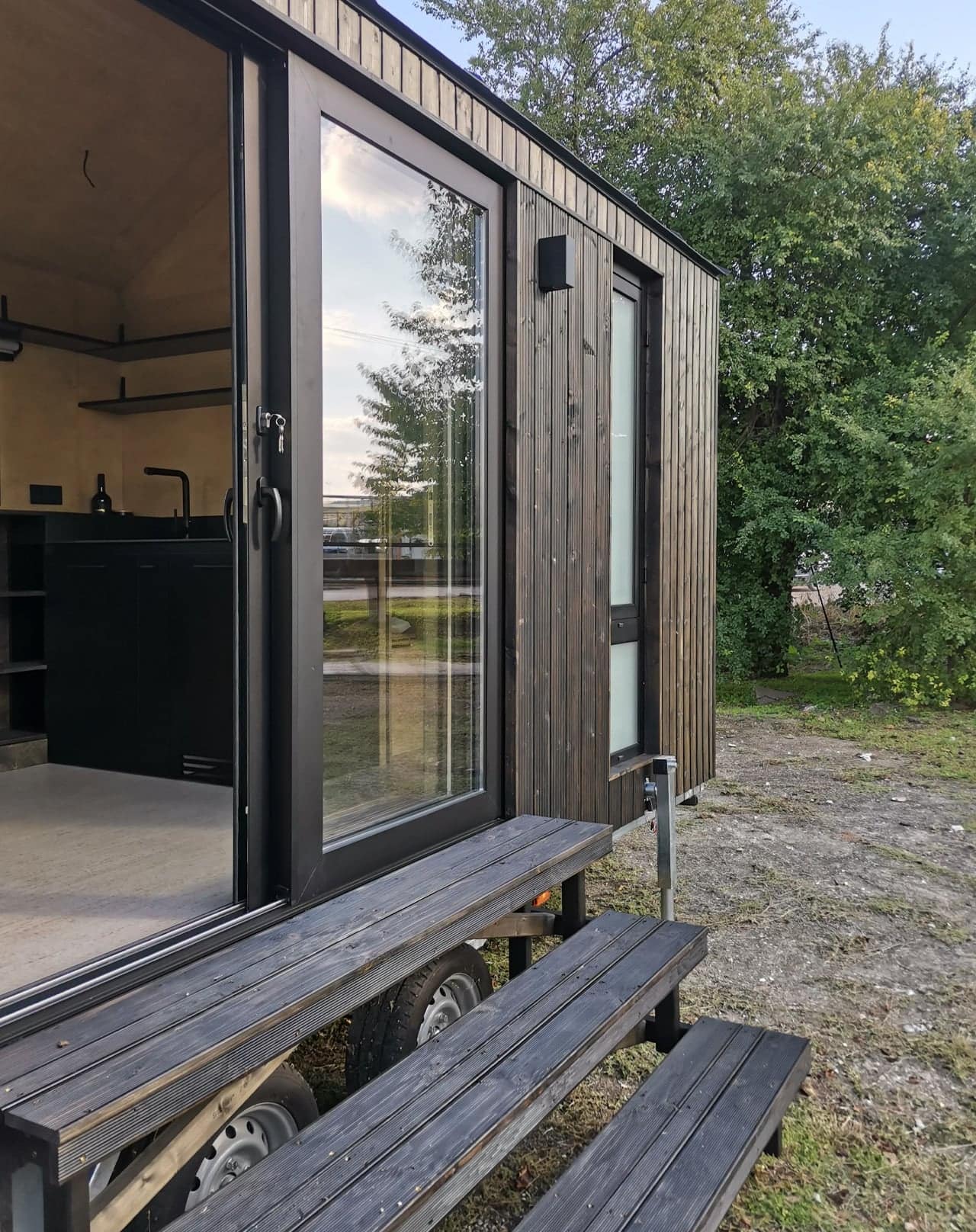 Sliding glass entry door of the Shoji tiny house with timber steps, showing the black kitchen and floating shelves visible inside