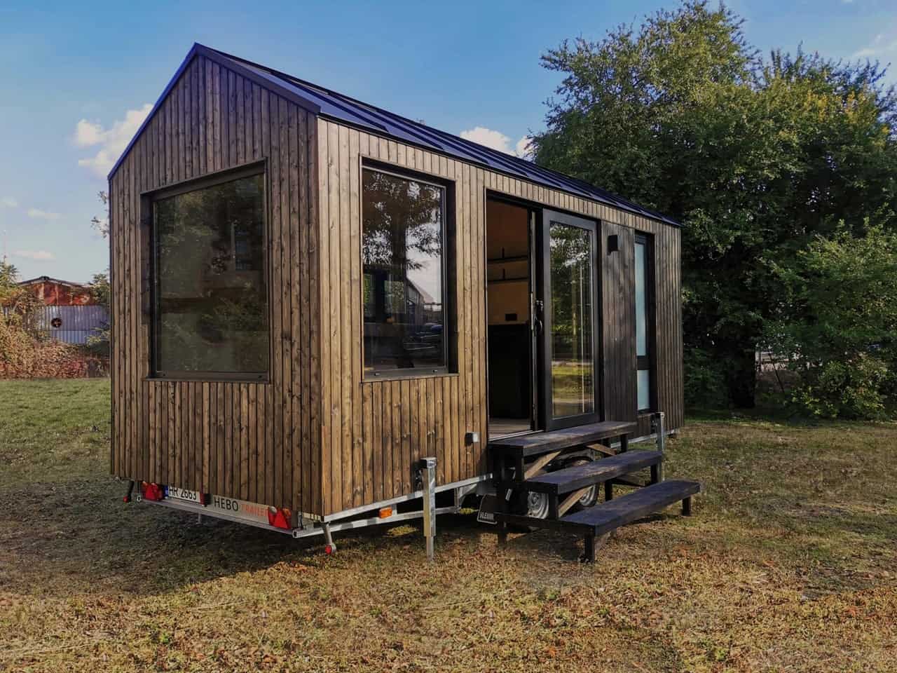 Shoji tiny house by Koleliba with dark vertical timber siding, black standing seam metal roof, corner picture window, and sliding glass door with timber steps