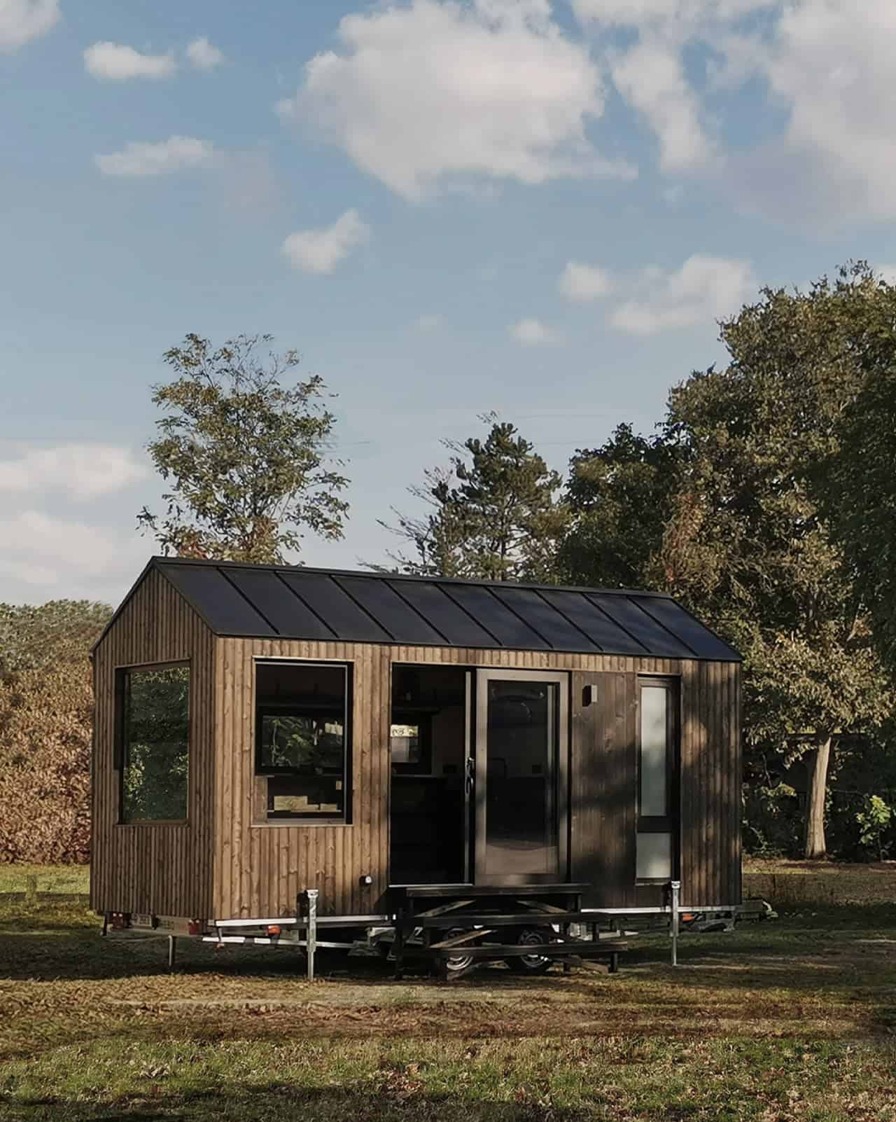 Shoji tiny house on a double-axle trailer with stabilizing legs down, sited in an open field with the black gable roof against blue sky