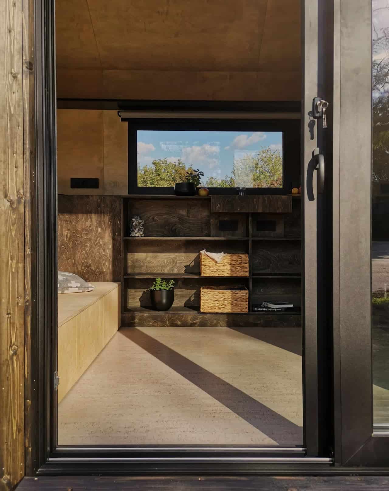 Interior of the Shoji tiny house seen through the open sliding door, with birch plywood walls, an open cubby storage wall, awning window, and plywood bench seating