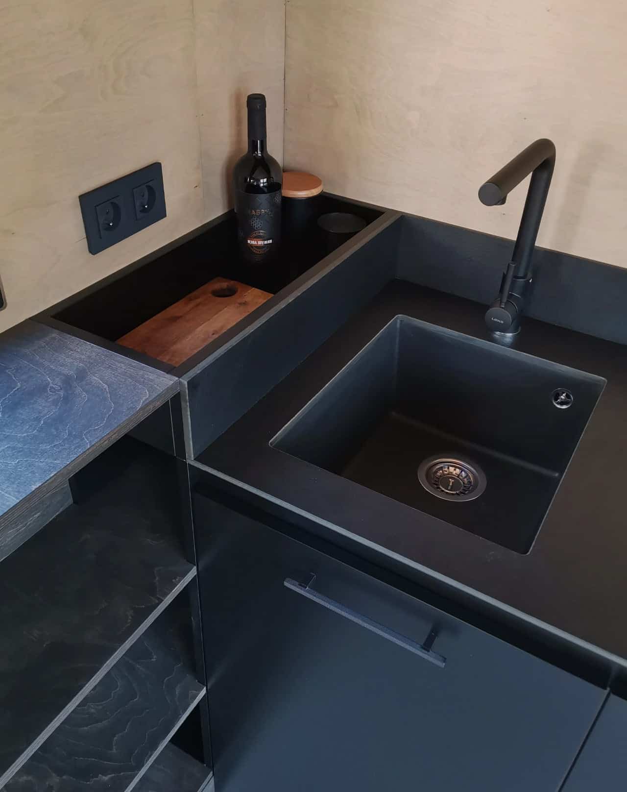 Close-up of the Shoji tiny house kitchen counter showing a black granite composite sink, black faucet, and a recessed timber cutting board set flush into the countertop