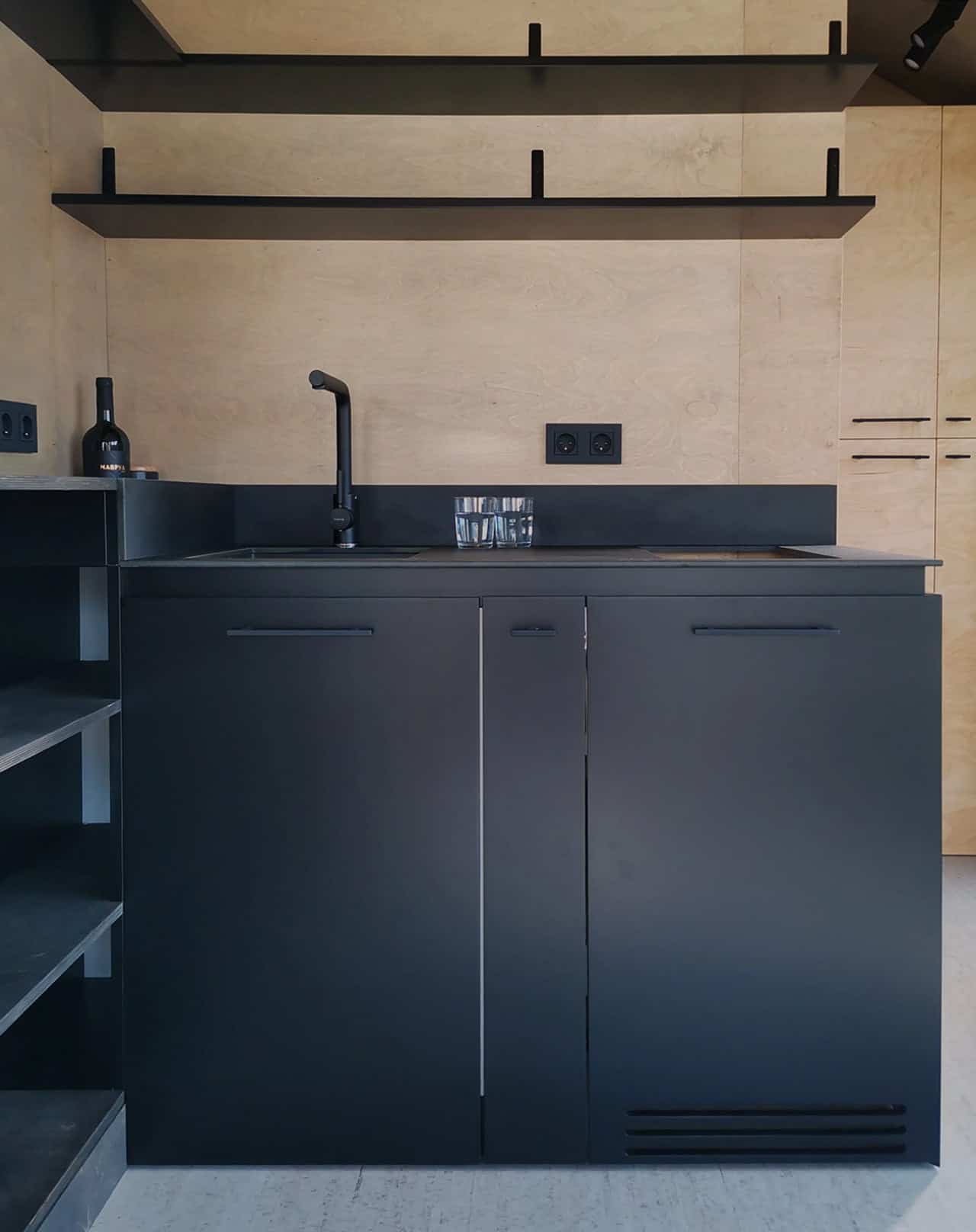 Matte black kitchen in the Shoji tiny house with black cabinetry, integrated cooktop, sink, and two slim black floating shelves against birch plywood walls